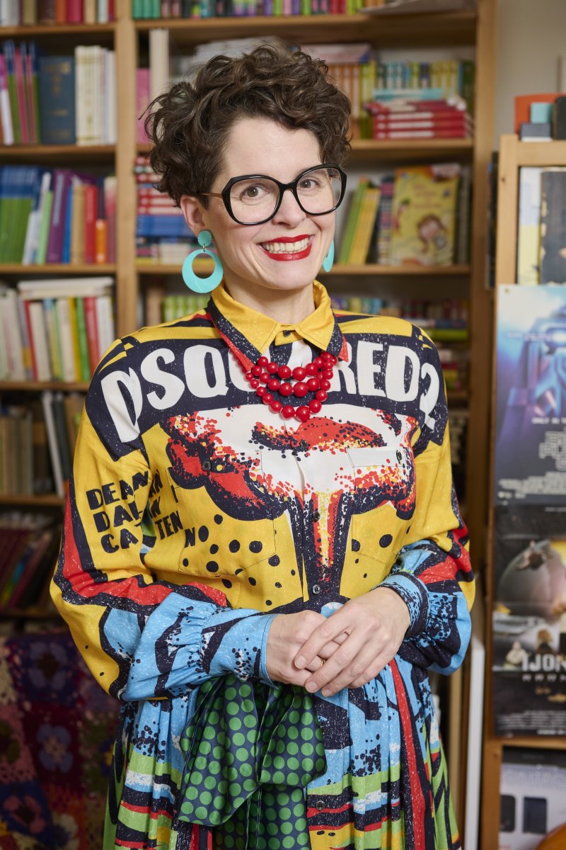 Frau mit Brille, buntem Hemd und roter Kette vor Bücherregal. Lächeln, Mode, Stil.