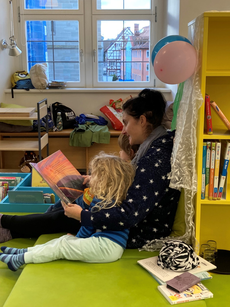 Mutter liest Kind in Bibliothek vor. Bücherregal, Fenster, Luftballons im Hintergrund. Leseförderung.