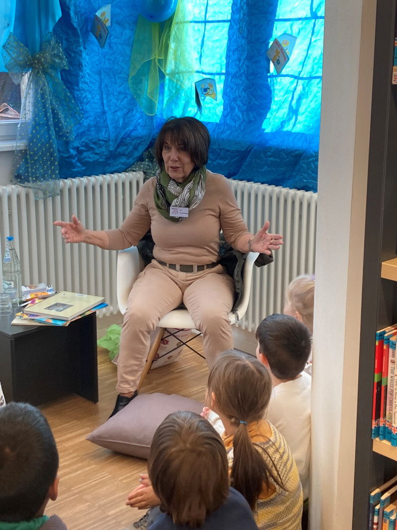 Frau liest Kindern vor. Vorlese-Event in Bibliothek. Kinder hören gespannt zu.
