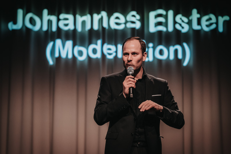 Johannes Elster, Moderator auf der Bühne mit Mikrofon. Bühnenbeleuchtung und Vorhang im Hintergrund.